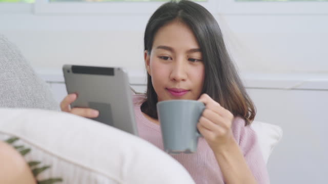 Schöne-attraktive-lächelnde-asiatische-Frau-mit-Tablet-hält-eine-warme-Tasse-Kaffee-oder-Tee,-während-auf-dem-Sofa-liegen,-wenn-im-Wohnzimmer-zu-Hause-entspannen.-Lifestyle-Frauen-zu-Hause-Konzept.
