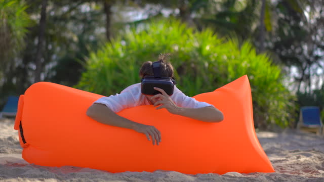 Young-man-on-an-inflatable-sofa-on-a-tropical-beach-uses-a-VR-glasses.-He-feels-like-he-is-swimming-in-a-sea-watching-lots-of-tropical-fishes.-Concept-of-modern-technologies-that-can-make-you-feel-like-you-are-somewhere-else