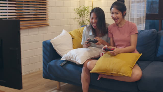 Lesbian-lgbt-women-couple-play-games-at-home,-Asian-female-using-joystick-having-funny-happy-moment-together-on-sofa-in-living-room-in-night.-Young-lover-football-fan,-celebrate-holiday-concept.