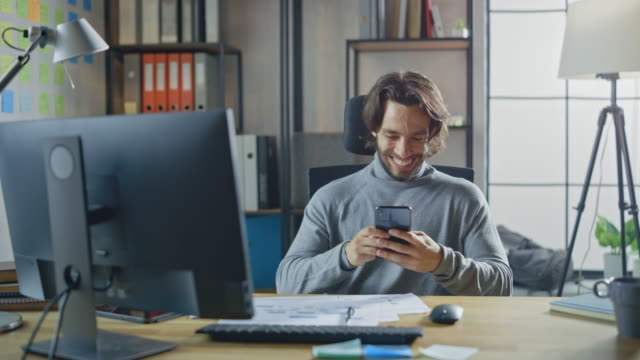Handsome-Long-Haired-Entrepreneur-Sitzt-an-seinem-Schreibtisch-im-Büro-arbeitet-auf-Desktop-Computer,-Arbeiten-mit-Dokumenten,-Graphen.-Lächelnd-verwendet-Smartphone,-Social-Media-App,-Schreiben-von-E-Mails,-Messaging