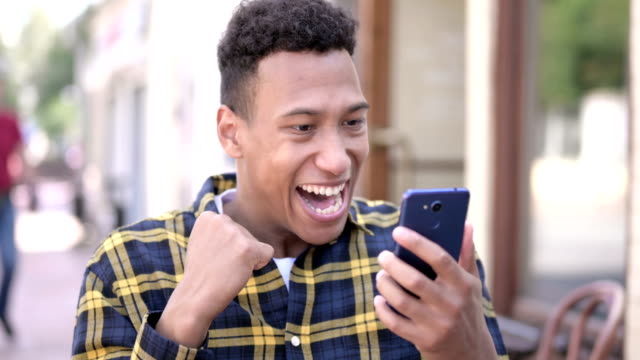 Hombre-africano-celebrando-el-éxito-en-línea-en-Smartphone,-al-aire-libre