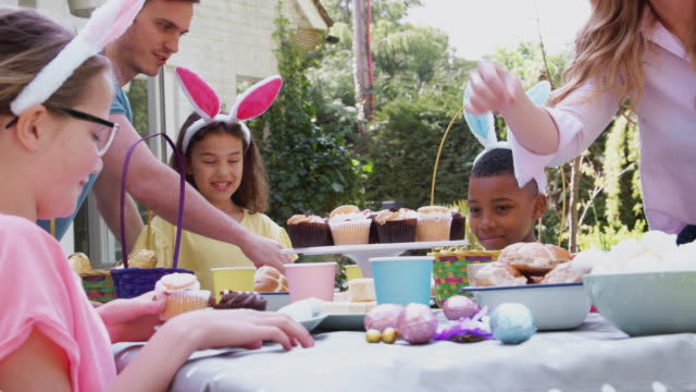 Grupo-de-niños-con-orejas-de-conejo-sentados-en-la-mesa-al-aire-libre-disfrutando-de-la-fiesta-de-Pascua-con-los-padres---tiro-a-cámara-lenta