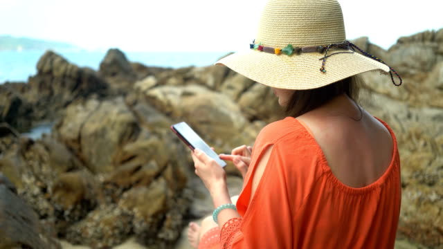 joven-hermosa-mujer-usando-el-teléfono-para-comunicarse-en-las-redes-sociales-y-sentarse-en-una-piedra-cerca-del-mar