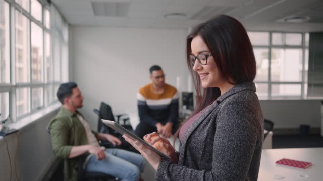 Portrait-of-young-confident-businesswoman-using-tablet-computer-smiling-and-looking-at-camera-standing-in-front-of-her-colleague-talking-in-the-background