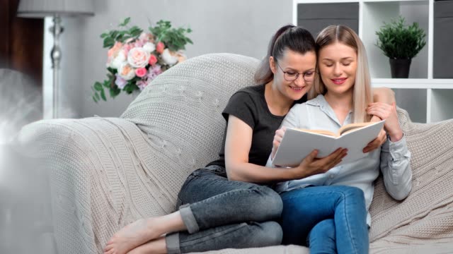 Adorable-happy-same-sex-female-couple-smiling-reading-book-together-relaxing-on-comfortable-couch