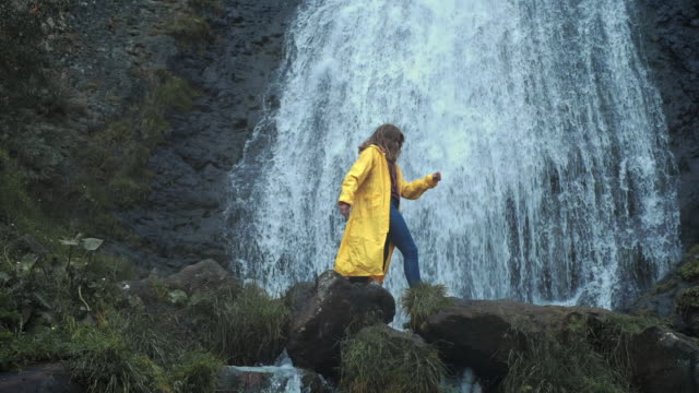 Young-girl-traveler-hiker-in-a-yellow-raincoat-walks-to-a-waterfall-in-the-highlands,-raises-his-hands-up,-enjoys-nature-and-life.-Traveling-in-the-mountains,-adventure-in-trip.-Lifestyle-concept