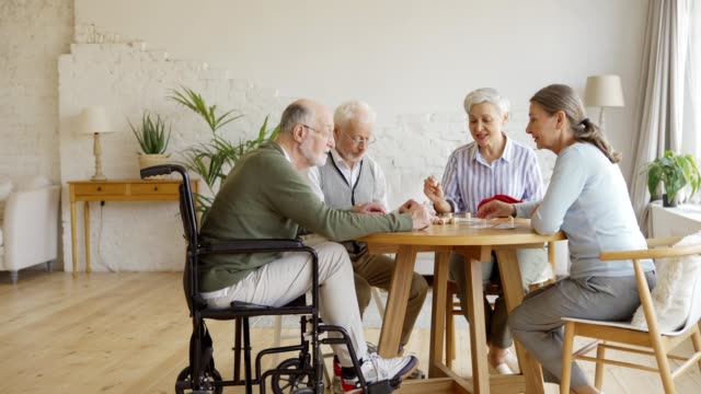 Seguimiento-de-la-toma-de-grupo-de-cuatro-ancianos-jubilados,-dos-hombres-y-dos-mujeres,-sentados-a-la-mesa-y-jugando-al-bingo-juntos-en-la-vida-asistida-en-casa-asistida