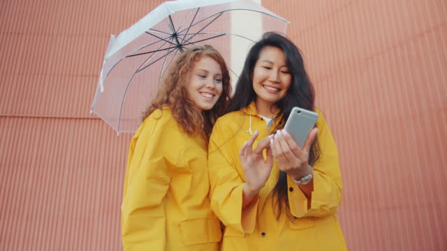 Fröhliche-junge-Frauen-in-Regenmänteln-mit-Smartphone-im-Freien-unter-Regenschirm