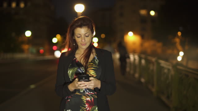 Nette-attraktive-stilvolle-kaukasische-moderne-Frau-trägt-Blumenkleid,-schwarze-Jacke-und-rote-Haare-zu-Fuß-durch-die-Straße-und-schreiben-eine-SMS-auf-ihrem-Smartphone-bei-Nacht.-Paris-4K-UHD.