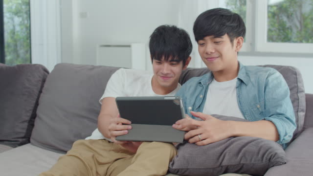 Joven-pareja-Gay-usando-tableta-en-casa.-Los-hombres-LGBTQ-asiáticos-feliz-relajarse-divertido-utilizando-la-tecnología-viendo-películas-en-Internet-juntos-mientras-se-acuesta-sofá-en-el-concepto-de-sala-de-estar.-Disparo-a-cámara-lenta.