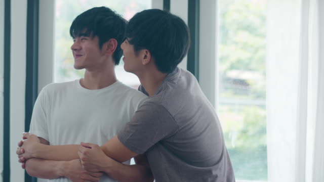 Pareja-gay-asiática-de-pie-y-abrazando-cerca-de-la-ventana-en-casa.-Jóvenes-asiáticos-LGBTQ+-hombres-besando-feliz-relax-descanso-juntos-pasar-tiempo-romántico-en-la-sala-de-estar-en-la-casa-moderna-en-el-concepto-de-la-mañana.