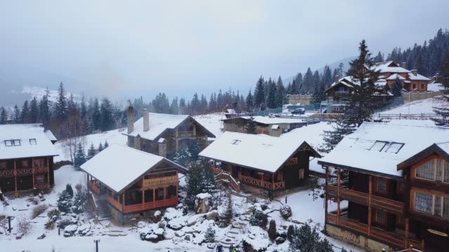Aire-de-cabañas-de-madera-en-pueblo-de-montaña-rodeado-de-bosque-de-coníferas.-Vista-de-drone-sin-chalets-cubiertos-de-nieve-en-la-estación-de-esquí.-Frío-día-de-invierno-helado-y-nevadas-en-las-montañas