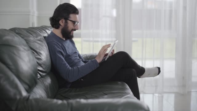 Side-view-of-bearded-Caucasian-man-in-eyeglasses-using-tablet-and-smiling.-Young-guy-sitting-on-comfortable-sofa-and-using-social-media.-Internet-addiction,-leisure-activity.