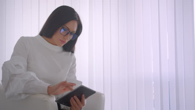 Closeup-side-view-portrait-of-young-pretty-caucasian-businesswoman-in-glasses-using-the-tablet-looking-at-camera-smiling-happily-sitting-in-the-armchair