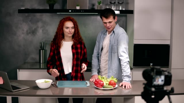 Young-couple-in-casual-cooking-together-and-recording-video-food-blog-on-camera-in-the-loft,-grey-kitchen-at-home.-Excited-welcome-their-followers,-looking-to-the-shooting-camera-on-foreground