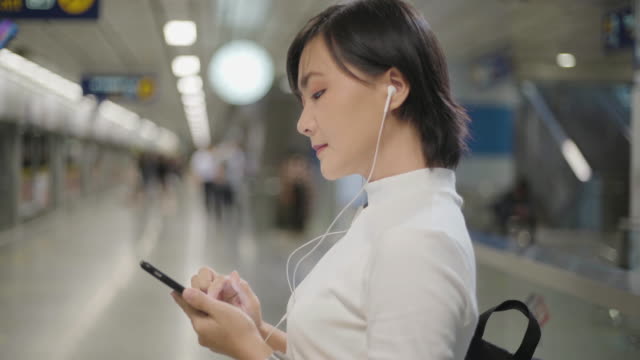 Porträt-der-asiatischen-Frau-mit-Kopfhörer-hören-Musik-und-mit-Dem-Smartphone-zum-Chatten-mit-Freunden-oder-Surfen-während-des-Wartens-auf-einen-Zug.-Technologie-im-Alltag-und-auf-Reisen.