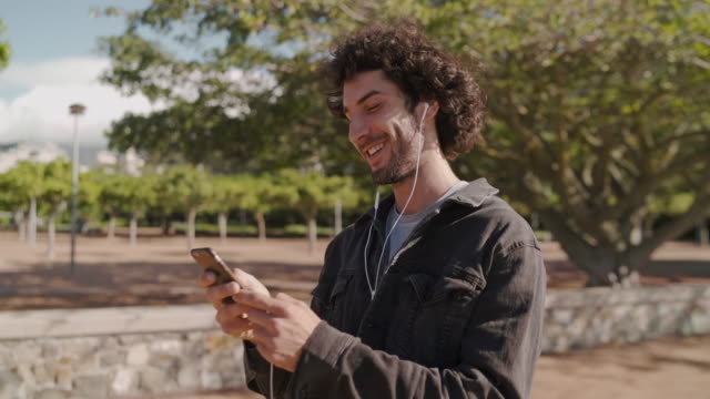 Fröhlicher-junger-stylischer-Mann-mit-Kopfhörern-tragen-Jeansjacke-mit-Smartphone-zum-Plaudern-mit-Freunden-während-des-Spaziergangs-im-Park