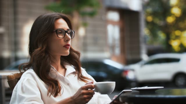Business-girl-in-glasses-and-white-shirt.-She-sitting-at-table-in-roadside-cafe.-Browsing-news-on-mobile-phone-and-drinking-coffee.-Slow-motion