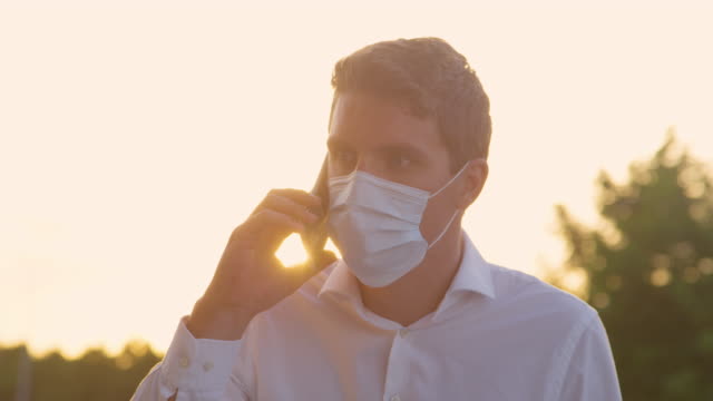 CLOSE-UP:-Man-wearing-a-facemask-gets-annoyed-while-talking-on-phone-at-sunset.