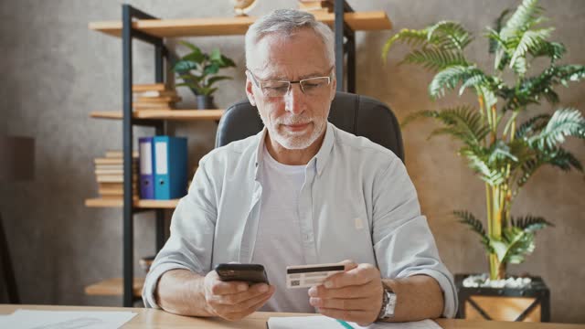 Senior-Geschäftsmann-hält-Kreditkarte-und-gibt-seine-Nummer-in-Smartphone,-Online-Shopping-sitzen-am-Schreibtisch