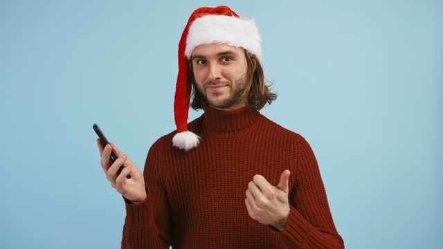 Guy-in-Santa-Hut-mit-Smartphone,-zeigt-seinen-schwarzen-Bildschirm,-lächelnd-und-zeigt-auf-sie,-hebt-Daumen-nach-oben,-blauen-Hintergrund