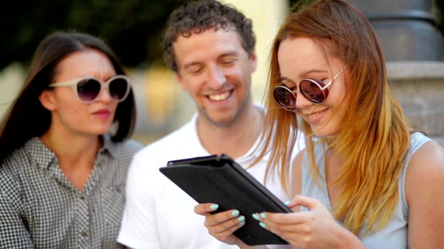 Group-of-Students-with-Tablet-Hanging-out.-Girl-in-Light-Blue-Dress-is-Reading-Something,-Her-Female-Friend-and-Boyfriend-Listen-Sitting-Outdoors-During-Warm-Sunny-Day-in-the-City