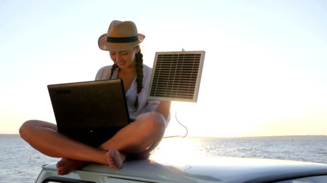 girl-in-vacation-sits-on-roof-car-with-solar-array-charges-laptop-in-backlight,-female-sitting-on-vintage-car-with-computer