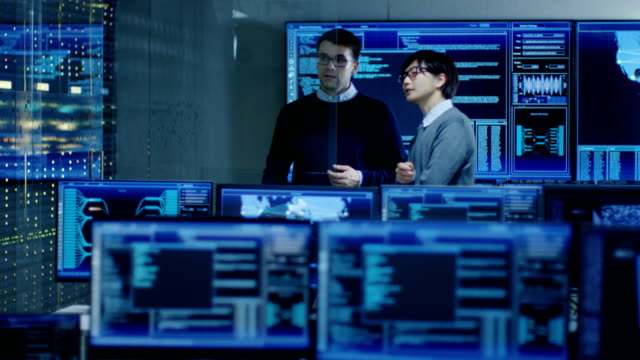 In-the-System-Control-Room-Project-Manage-and-IT-Engineer-Have-Discussion,-they're-surrounded-by-Multiple-Monitors-with-Graphics.-They-Work-in-a-Data-Center-on-Data-Mining,-AI-and-Neural-Networking.
