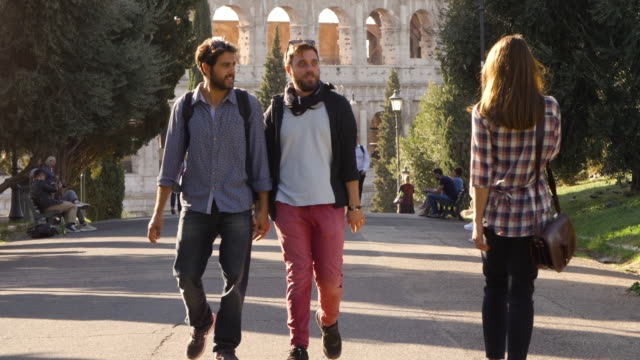 Young-lovely-gay-couple-tourists-walk-in-park-road-with-trees-colosseum-in-background-in-rome-at-sunset-holding-hands.-Boyfriend-gets-jealous-when-beautiful-girl-walks-by-glancing.-Slow-motion