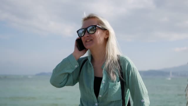 Beautiful-slim-woman-with-long-blonde-hair-and-green-shirt-standing-and-talking-on-phone-over-background-sea