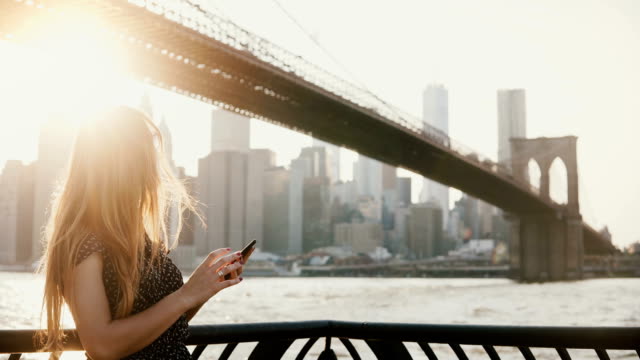 Schönes-Mädchen-mit-langen-Haaren-in-Sonnenbrille-mit-Smartphone-app-am-Sonnenuntergang-Fluss-Kai-in-der-Nähe-von-Brooklyn-Bridge-New-York-4K