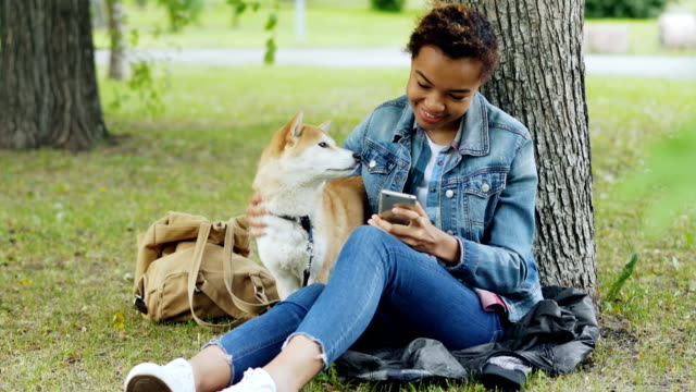 Sociable-girl-is-using-smartphone-texting-friends-and-stroking-her-adorable-puppy-while-resting-in-park-at-weekend.-Modern-technology,-people-and-animals-concept.