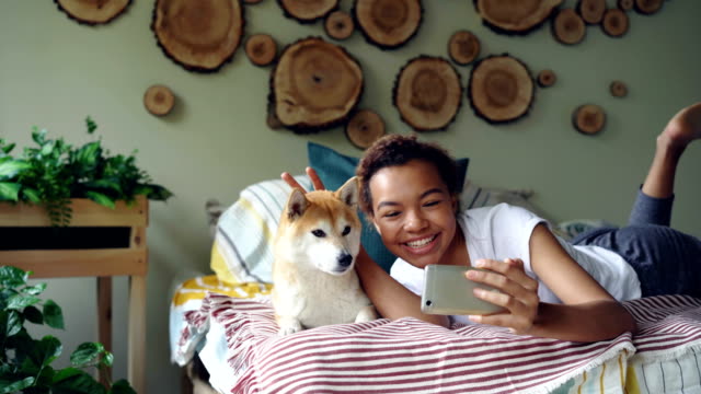 Fröhliches-Mädchen-nimmt-Selfie-mit-Haustier-posiert-mit-schöner-Hund-auf-Bett,-Spaß-haben-und-lachen.-Modernes-Apartment-mit-schönen-Design-und-Möbel-ist-sichtbar.