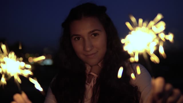 Eine-junge-Frau-mit-langen-dunklen-Haaren-hält-Feuerwerk-in-der-Nacht-im-Hintergrund-der-Stadt-und-ist-glücklich,-guten-Laune-zu-haben.-Slow-Motion.-Porträt.-4K