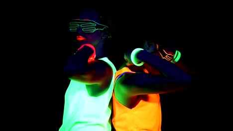 Women-with-UV-face-paint,-glowing-bracelets,-glowing-glasses,-clothing-dancing-back-against-each-other-in-front-of-camera,-Half-body-shot.-Caucasian-and-asian-woman.-Women.