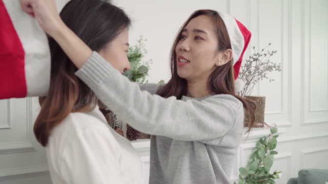 Lesbian-Asian-couple-giving-and-wear-christmas-hat-to-each-other-in-her-living-room-at-home-in-Christmas-Festival.-Lifestyle-lgbt-women-happy-celebrate-Christmas-and-New-year-concept.
