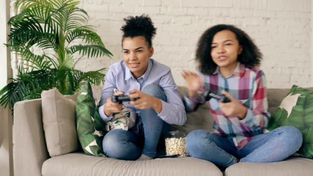 Zwei-gemischte-Rennen-lockige-Mädchen-Freunde-sitzen-auf-der-Couch-spielen-Konsole-Computerspiele-mit-Gamepad-und-Spaß-zu-Hause