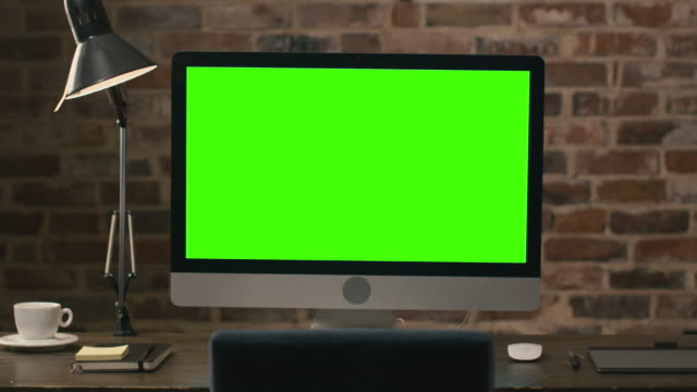 Aufnahmen-eines-Computermonitors-mit-grünem-Bildschirm-auf-einem-Tisch-neben-einem-Tablet,-Lampe,-Kaffeetasse,-Notebook-und-Maus-in-einem-Loft.