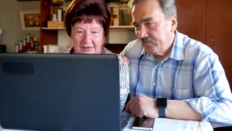 An-elderly-couple-is-sitting-at-home-at-the-laptop.-A-woman-reads-news,-a-man-with-a-mustache-sits-next-to-him-and-talks