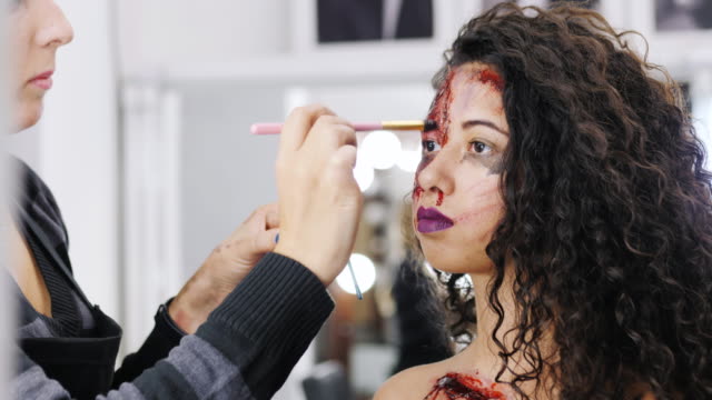 Make-up-artist-make-the-girl-halloween-make-upin-studio.Halloween-face-art.Woman-applies-on-professional-greasepaint-on-the-face-of-spanish-girl.War-paint-with-blood,-scars-and-wounds.-4k