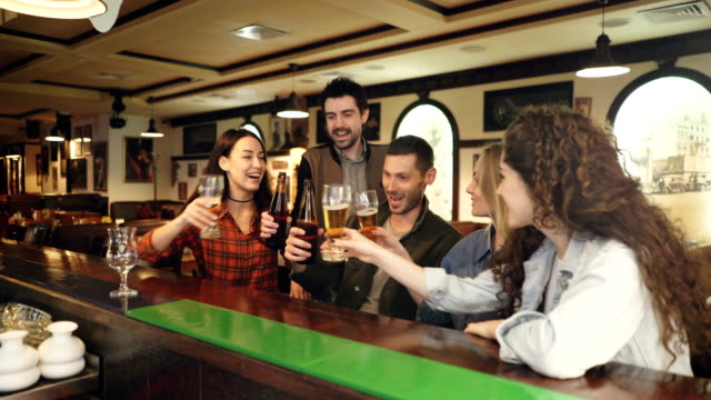 Young-people-are-celebrating-something-over-beer-in-bar.-They-are-proposing-toast,-clanging-glasses-and-bottles,-chatting-and-laughing.-Celebration-happiness-concept.