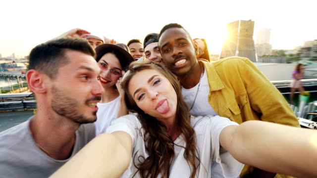 Attraktive-junge-Frau-nimmt-Selfie-mit-Freunden-auf-Dach,-Mädchen-Kamera-hält-und-posieren,-während-ihre-Kollegen-lustig-machen-Spaß-haben-Gesichter-und-Gesten.