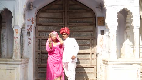Lovely-Indian-couple-taking-camera-selfie-on-mobile-phone-photography-in-front-of-their-house-with-traditional-architecture-gateway-house-exterior