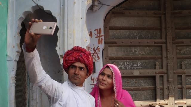 Dolly-in-to-Couple-taking-camera-selfie-on-mobile-phone-photography-in-front-of-their-house-with-traditional-architecture