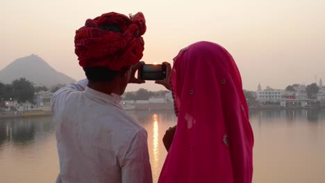 Pan-izquierda-a-pareja-en-vestido-indio-tomar-fotos-del-atardecer-en-un-lago-sagrado-en-Rajasthan
