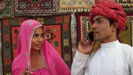 Tilt-up-to-Indian-couple-in-traditional-clothes-busy-on-a-phone-call-with-traditional-carpets-hanging-in-the-backdrop