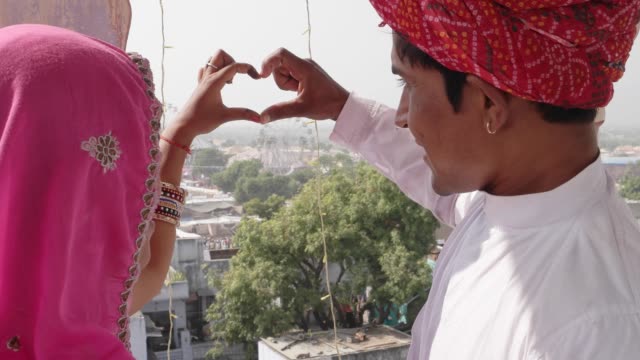 Indian-couple-in-traditional-dress-make-heart-symbol-with-fingers-and-hand,-looking-down-from-a-vantage-point-at-Pushkar-Mela-festival-in-Rajasthan,-India