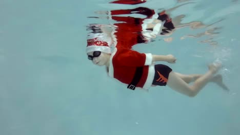 Ein-kleiner-Junge-in-einem-Anzug-und-Hut-Weihnachtsmann-schwimmt-unter-Wasser-im-Pool-mit-Brille-zum-Schwimmen-und-in-die-Kamera-schaut.-Slow-Motion.-Seitenansicht.-RAW-Video.-Fotografieren-unter-Wasser.