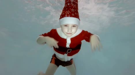 Mom-helps-a-little-boy-dressed-as-Santa-Claus-to-dive-under-the-water-in-the-children's-pool.-The-boy-swims,-smiles-and-looks-at-the-camera.-Slow-motion.