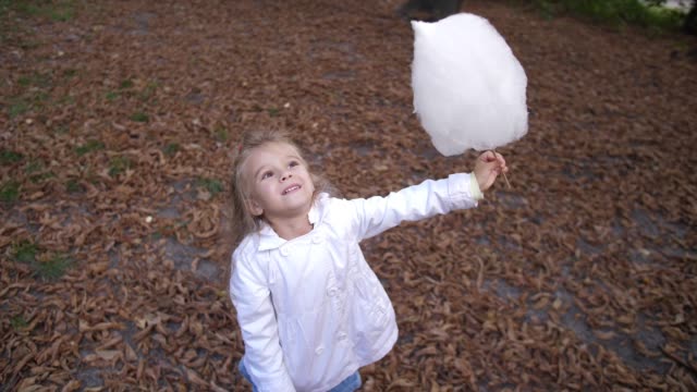 Little-blonde-girl-is-eating-sweet-cotton-candy-in-the-city-park.-Beautiful-little-girl-eating-candy-floss.-Child-eating-cotton-candy-green-forest-at-the-backgorund.-4k.-Slow-Motion